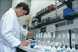 Ein Mensch in weißem Kittel steht in einem Chemielabor vor einem Tisch mit zahlreichen Kolben, Flaschen, Bechergläsern und Tinkturen, und notiert etwas auf einem Klemmbrett..