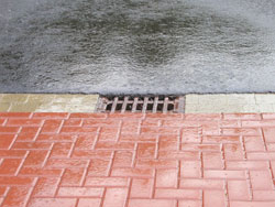 Regenwasser-Gully in der Rinne zwischen rot gepflastertem Fußweg und asphaltierter Straße bei Regen.