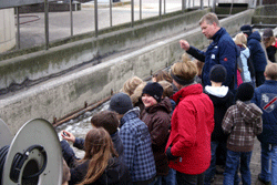 Eine Gruppe Schüler*innen steht vor einem Schacht aus Beton; ein Mitarbeiter des Klärwerkes erläutert etwas dazu.