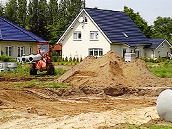Kanalverlegungs-Baustelle in einem Einfamilienhaus-Neubaugebiet. Ein Radlader transportiert Rohrteile neben aufgeschüttetem Sandaushub.