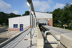 Ein langgestreckter Schacht, abgegrenzt durch eine Mauer und weitere technische Einrichtungen, führt auf das Betriebsgebäude zu.