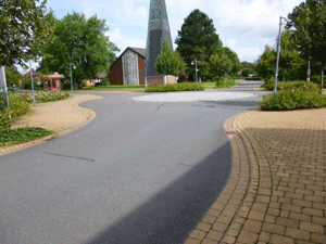 Ein Teil der Hauptstraße mit daran anschließendem Kreisverkehr, im Hintergrund die modern anmutende Kirche mit dem separat stehenden Turm.