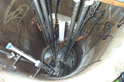 Blick senkrecht herunter in einen Pumpenschacht. Am Rand des Schachtes verläuft eine Leiter und mehrere Rohrleitungen.