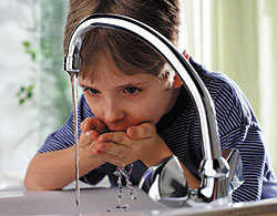 Aus einem Wasserhahn fließt Wasser, dahinter steht ein Kind, das aus den gefüllten Händen Wasser trinkt