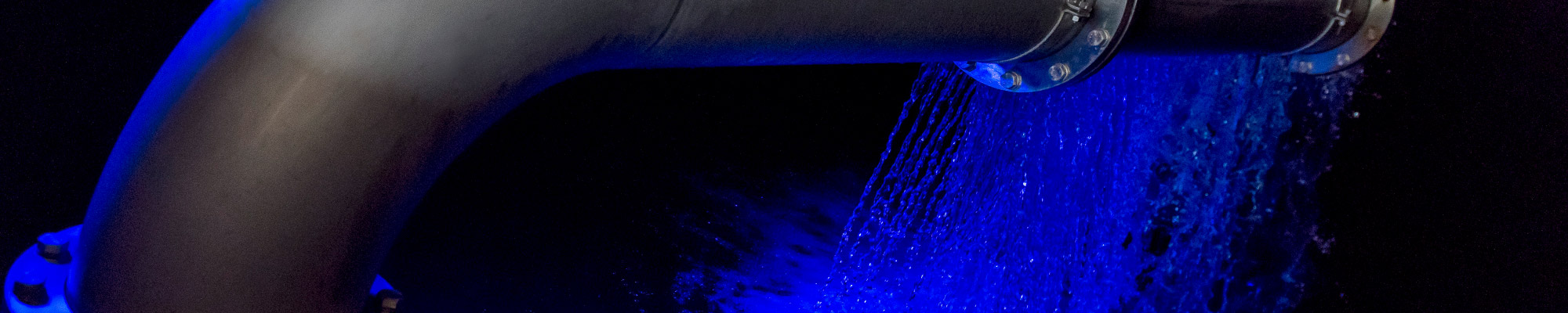 Detail einer Wasserrohrleitung, dahinter ein sprudelnder Wasserschwall, alles in blaues Licht getaucht.