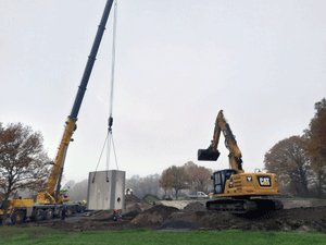 Kanal-Baustelle mit Aushub, rechts im Bild ein Raupenbagger, links hebt ein Kranwagen den Betonblock des Drossel Bauwerks in den Schacht.