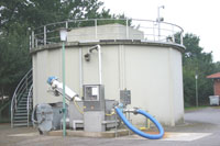 Der zylinderförmige Fäkalschlammspeicher, auf dem Dach von einem Geländer umgeben; davor die Annahmestation, ähnlich einer Tanksäule mit einem großen blauen Schlauch.