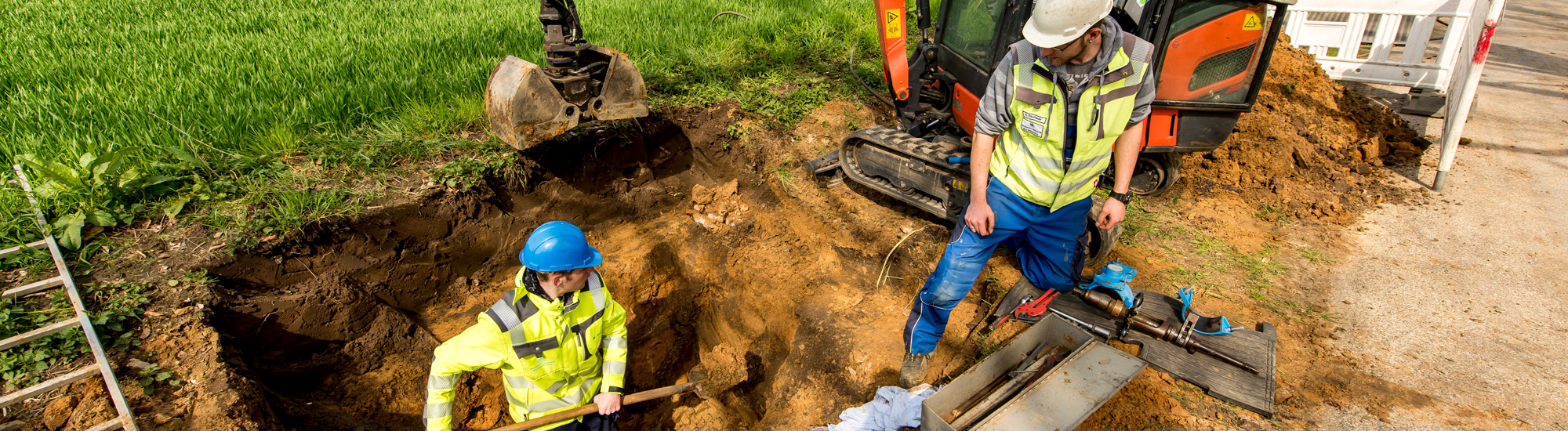 Arbeiter mit Schaufel in einem ausgehobenen Schacht; daneben ein weiterer Arbeiter, der in den Schacht schaut; im Hintergrund ein Kleinbagger