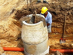 Arbeiter in einer ausgehoben Kanalgrube; ein Betonrohr wird neben ihm von einem Kran abgesetzt.