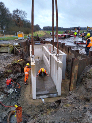 Ein großes Betonfertigteil wird von einem Kran in den vorbereiteten Schacht gehoben, in dem sich einige Bauarbeiter befinden; im Hintergrund ein vierachsiger Tieflader.