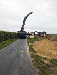 Sandaushub neben einem Landwirtschaftsweg. Auf der Straße steht ein Kranwagen und hebt das zylinderförmige Zwischenpumpwerk; im Hintergrund der Bagger, der den Aushub durchführt.