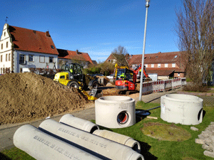 Kanal-Baustelle mit Radlader und Kleinbagger, ein großer Haufen Aushub; im Vordergrund mehrere Betonrohrteile.