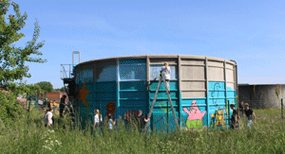 Mehrere Schüler*innen stehen – zum Teil auf Leitern – an der Außenwand eines kreisrunden Schmutzwasserspeichers und bemalen diesen.