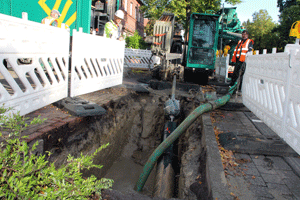 Ein ausgehobene Schacht neben einer Straße mit Absperr parken abgeteilt. An dem einen Ende steht das spezielle Baufahrzeug für das Lenkbohrverfahren. Von dort ragt ein Schlauch in den Schacht..