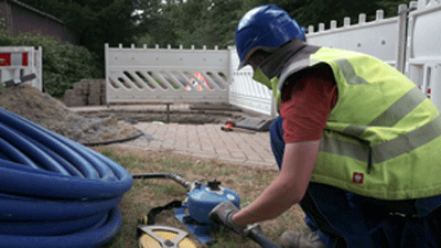 Ein Bauarbeiter in gelber Weste hockt neben einer Rohrleitung und hantiert mit einem technischen Gerät.