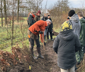 Förster mit Spaten auf Freifläche und Jugendliche, denen etwas erklärt wird.