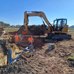Straßenbaustelle, ein Bagger hebt einen Schacht aus, durch den teilweise bereits ein Rohr verläuft. Einige Bauarbeiter bewegen sich drum herum mit verschiedenen Baugeräten.