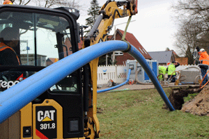 Von einem Baufahrzeug aus verläuft eine lange blaue Rohrleitung auf einen vorbereiteten Schacht zu und in diesen hinein.