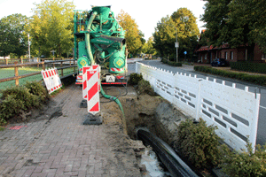 Ein teilweise geöffneter Schacht neben einer Straße, abgeteilt mit Absperr parken. In dem Schacht laufen mehrere Rohrleitungen entlang; aus einem Baufahrzeug ähnlich einem Betonmischer ragt ein Rohr in den Schacht, aus dem Wasser fließt.