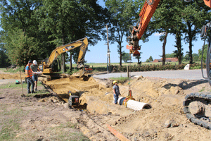 Ausgehobene Grube einer Kanal-Baustelle. In der Grube steht ein Bauarbeiter und nimmt ein Betonrohr entgegen, das von einem Kran herabgelassen wird. Daneben ist ein Bagger zu sehen und ein weiterer Bauarbeiter, der mit einer Fernbedienung den Kran steuert.