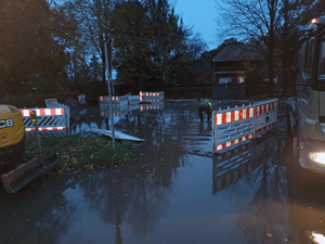 Überschwemmte Straße, nach einem Wasserrohrbruch, rot-weiße Balken grenzen die Fläche ab, Arbeiter in gelben Jacken und Baufahrzeuge hantieren auf der Fläche.