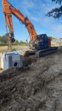 Ein Raupenbagger hebt den Betonblock des Drosselbauwerks in den bereits ausgeschachteten Kanal.