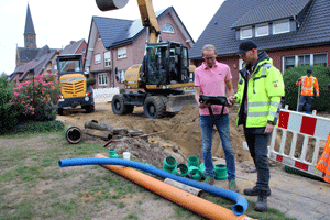 Kanalbaustelle mit Sandaushub, im Hintergrund ein Bagger und ein Radlader; vorne stehen zwei Menschen in Arbeitskleidung und diskutieren eine Unterlage.