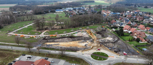 Luftaufnahme (Schrägansicht), einer Straße mit einem Kreisverkehr, dahinter eine Baustelle am Ortsrand