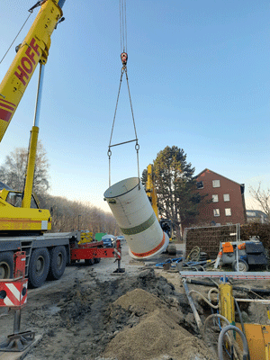 Ein Kranwagen hebt ein zylinderförmiges Teil des neuen Pumpwerkes in die ausgehobene Grube.