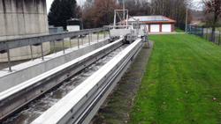 Ein langgestreckter Schacht, abgegrenzt durch eine Mauer und weitere technische Einrichtungen, führt neben einer Rasenfläche auf das Betriebsgebäude zu.