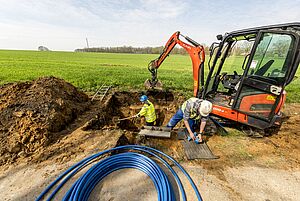 Aufgerollte blaue Schlauchleitungen in Nahaufnahme; dahinter steht ein Bauarbeiter in einer ausgehobenen Grube, ein weiterer kniet davor; neben der Grube steht ein Kleinbagger.