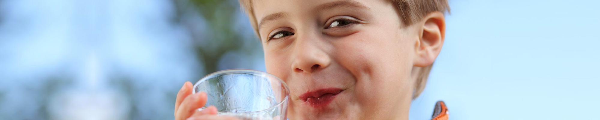 Ein kleiner Junge hält ein Glas Wasser in den Händen und grinst verschmitzt.