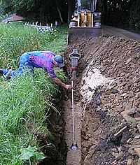 Rohrverlegungs-Baustelle; ein Arbeiter kniet neben einem ausgeschachteten Graben. Im Hintergrund steht ein Radlader mit Schaufel.