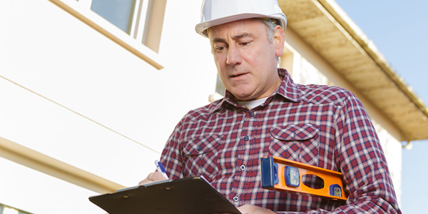 Ein Bauarbeiter mit kariertem Hemd und weißem Helm füllt ein Formular auf einem Klemmbrett aus, unter seinem Arm trägt er eine Wasserwaage. Im Hintergrund sieht man einen Rohbau.