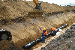 Eine ausgeschachtete Rinne neben einem Damm aus Kies und Sand; in der Rinne liegt eine schwarze Wasserrohrleitung. Daneben befinden sich zwei Arbeiter in orangener Kleidung mit Helm, darüber eine Baggerschaufel.