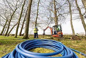 Aufgerollte blaue Schlauchleitungen in Nahaufnahme; im Hintergrund zwei Bauarbeiter und ein Kleinbagger zwischen Bäumen stehend