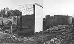 Eine halbkreisförmige Wand, aus der die Stahlarmierungen herausragen, bildet den halbfertigen Hochbehälter; im Hintergrund ein fertiger kreisförmiger Hochbehälter
