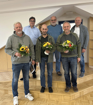 Sechs Männer stehen auf einer Treppe. In der ersten Reihe stehen die geehrten Mitarbeiter mit einem Blumenstrauß. In der zweiten Reihe stehen die drei Gratulierende.