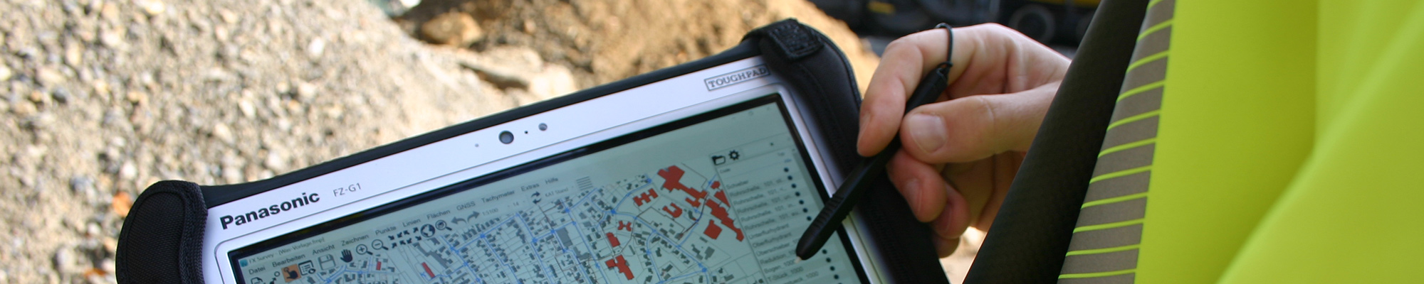 Ein Mensch in gelber Arbeitsjacke hält einen Tabletcomputer, auf dem eine Lageplan-Skizze zu sehen ist.