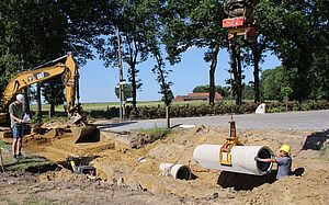 Ausgehobene Grube einer Kanal-Baustelle. In der Grube steht ein Bauarbeiter und nimmt ein Betonrohr entgegen, das von einem Kran herabgelassen wird. Daneben ist ein Bagger zu sehen und ein weiterer Bauarbeiter, der mit einer Fernbedienung den Kran steuert.