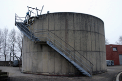 Der zylinderförmige Turm des Schlammsilos aus Betonplatten; am Rand führt eine Stahltreppe zum Dach des Silos.
